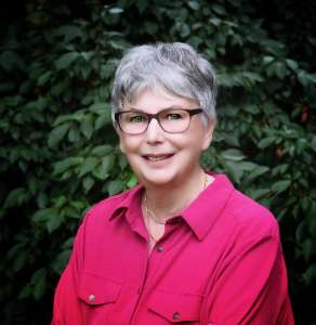 Writer Jackie Craven in a red shirt stands in front of dark green foliage.